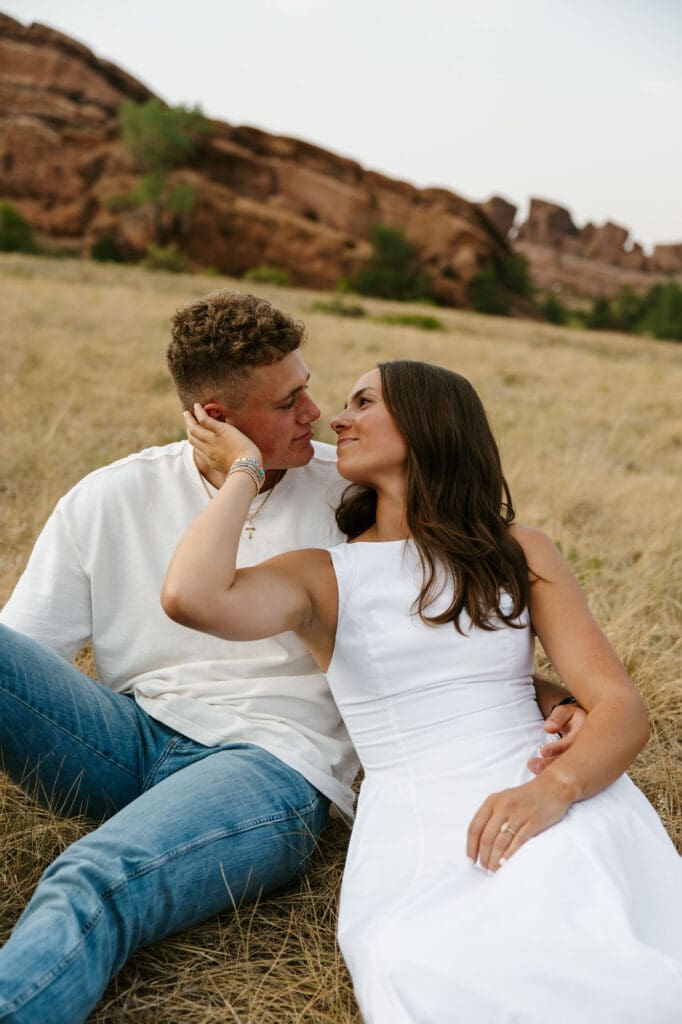 Jordan and Andrew sitting together in a grassy field with Red Rocks behind them during their engagement session